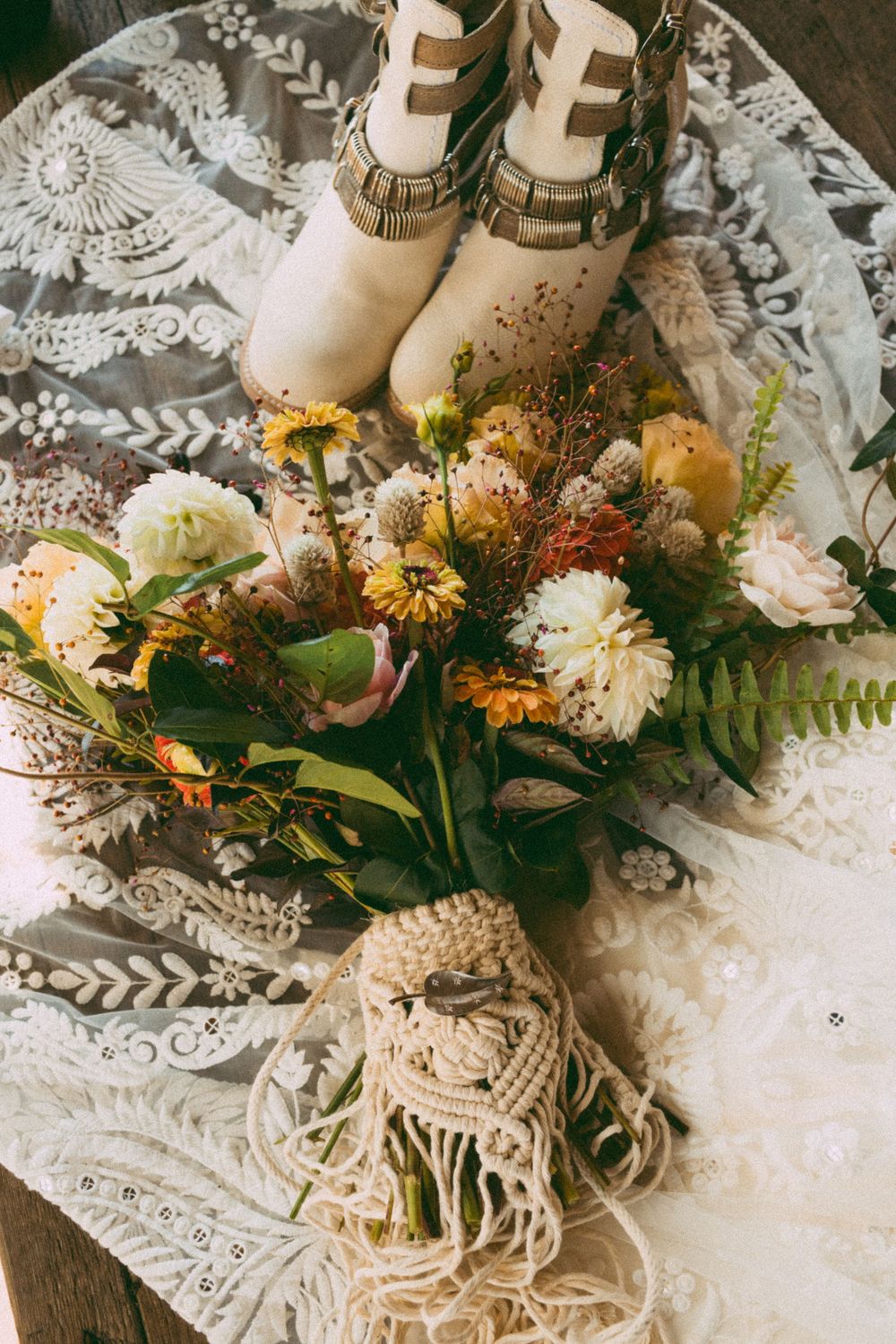 Rustic bridal bouquet with wildflowers and macrame wrap beside white boho boots on lace fabric.