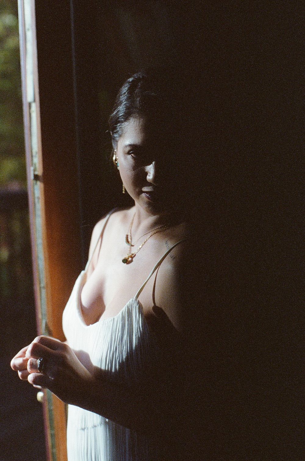 Woman in white dress standing in dramatic doorway light wearing gold necklace, artistic film photography portrait.
