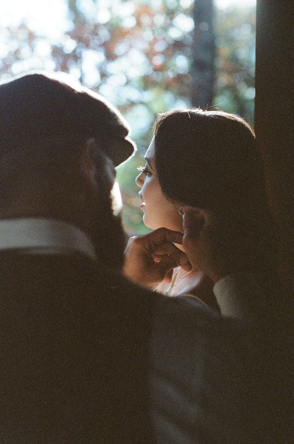 A couple shares an intimate moment in soft light, the woman gazing up tenderly at the bearded man near a window.
