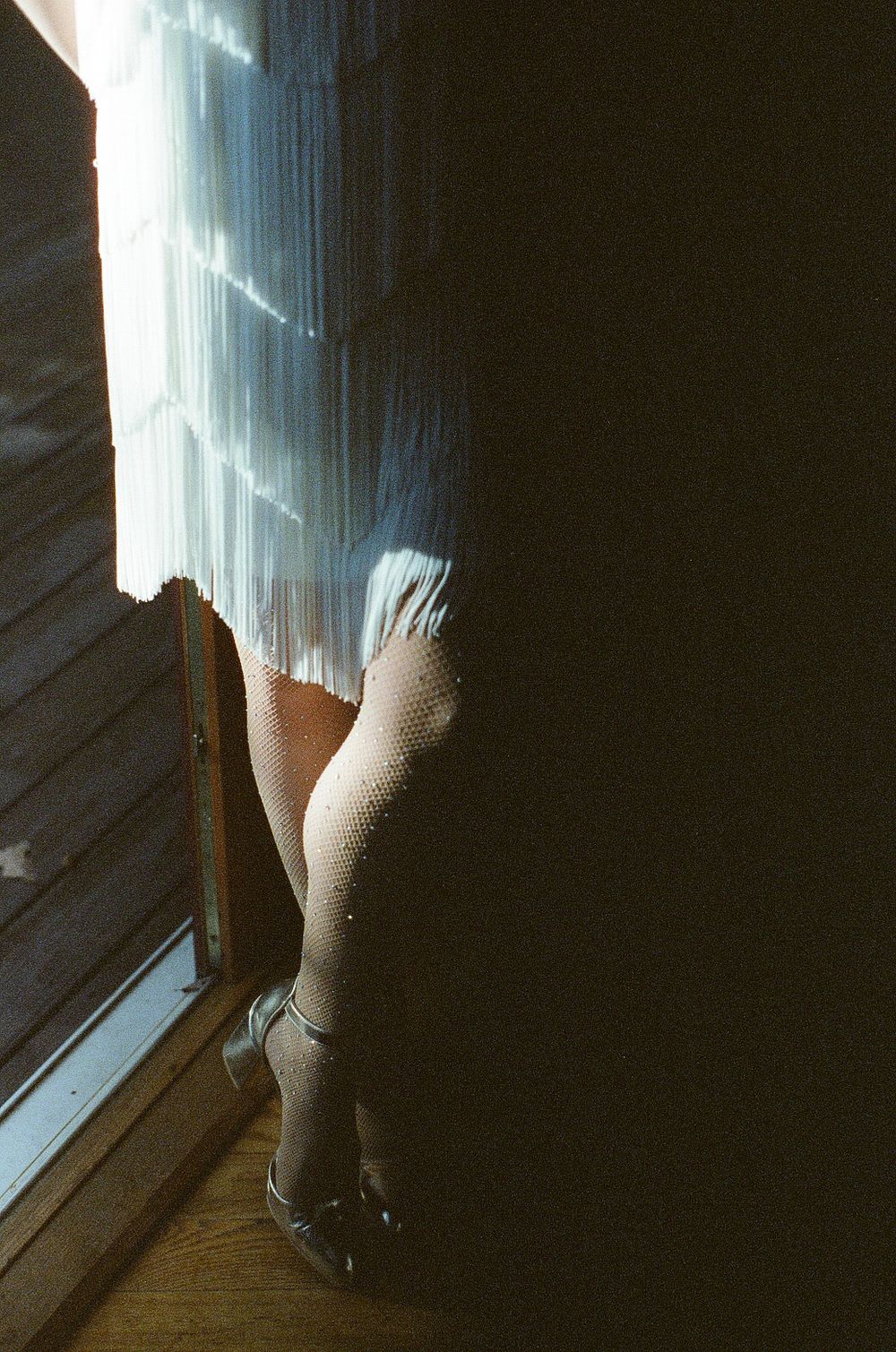 Person wearing fishnet stockings and heels standing by a sunlit window with a fringed skirt.