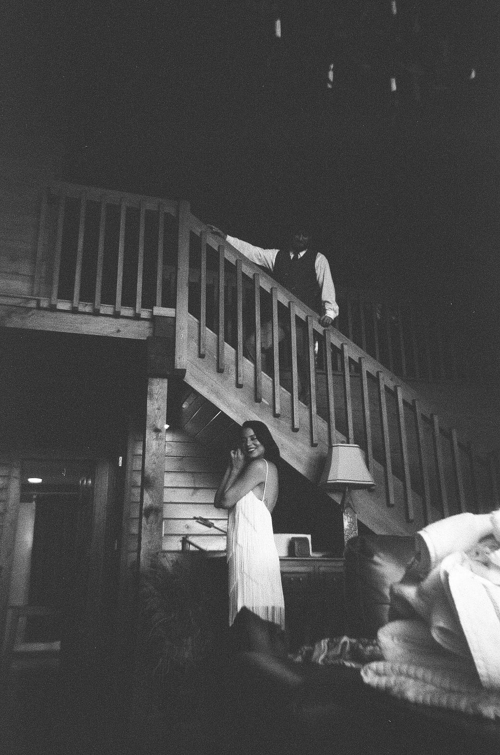 Black and white photo of a woman in white dress near wooden staircase with a man standing above in dimly lit interior.