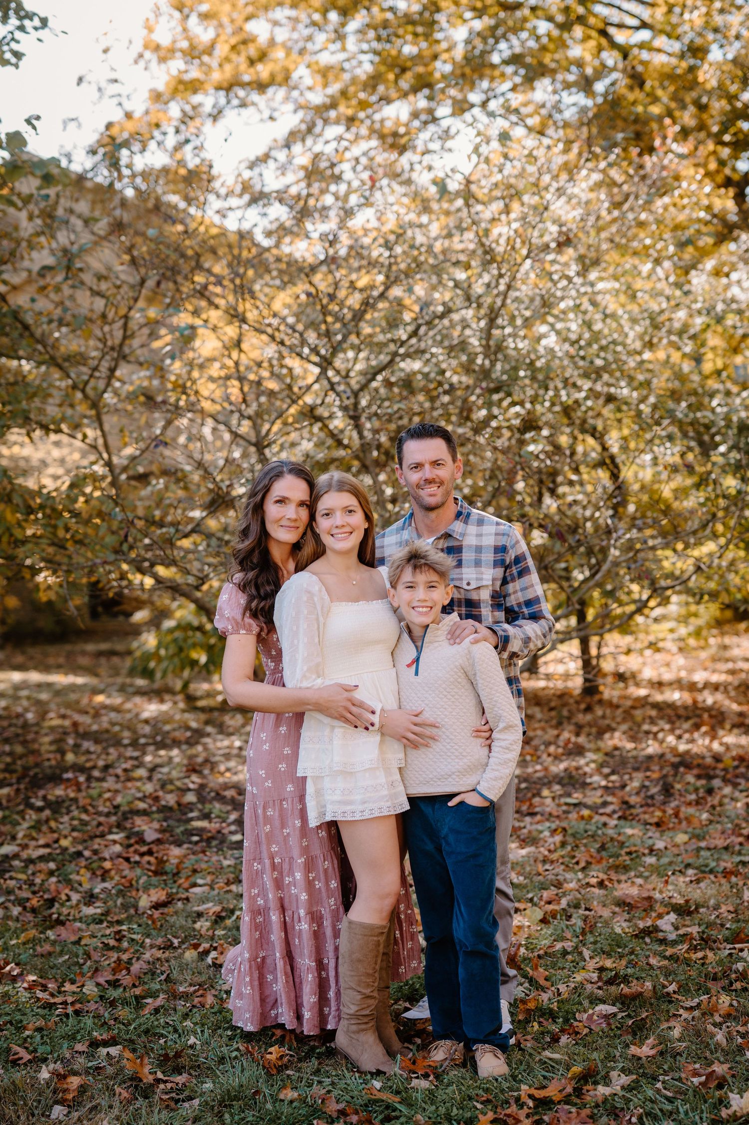 Four children pose outdoors in fall foliage, wearing coordinated outfits in green, burgundy, and blue for a family portrait.