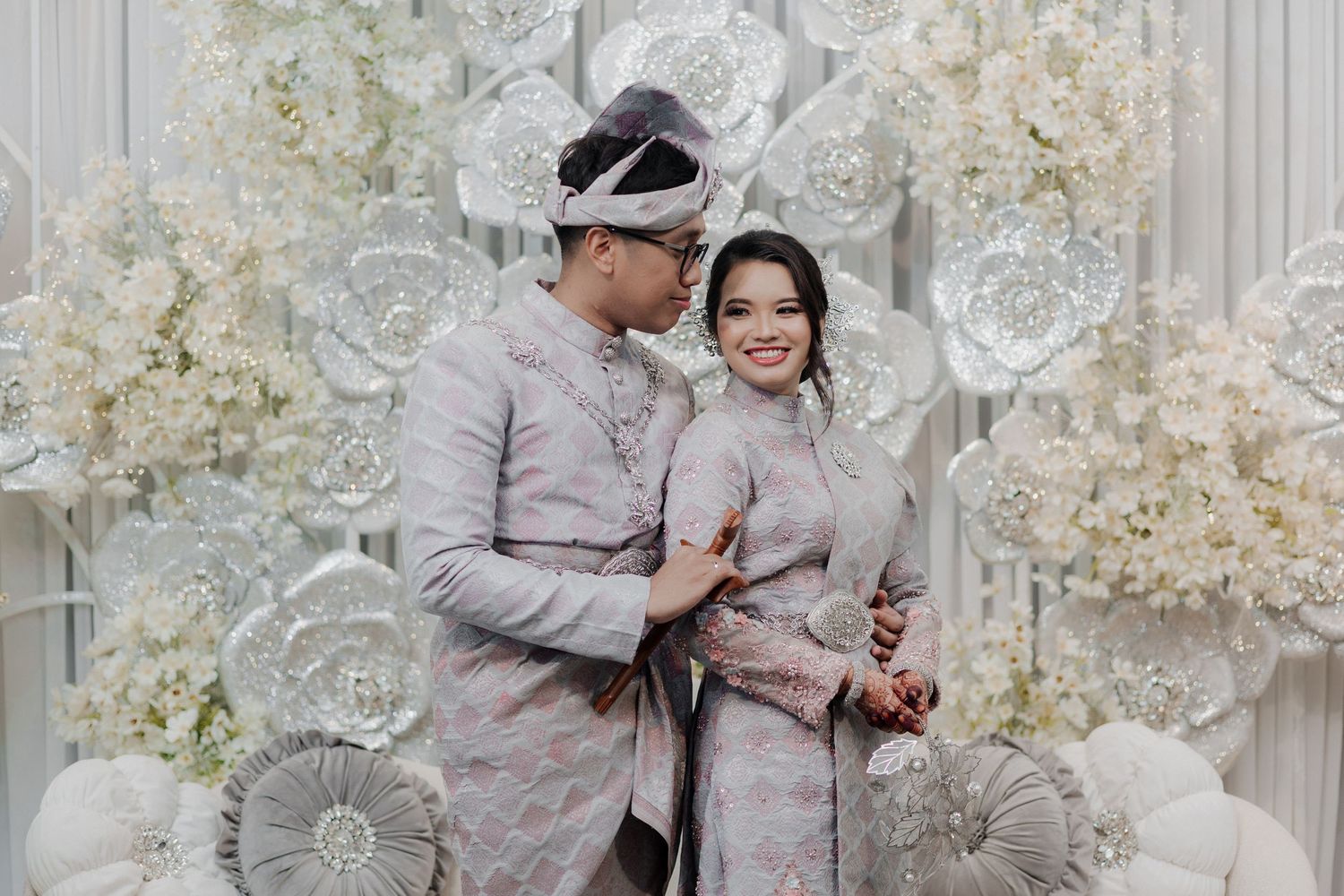 Couple in traditional Indonesian wedding attire smiling together against white floral backdrop.