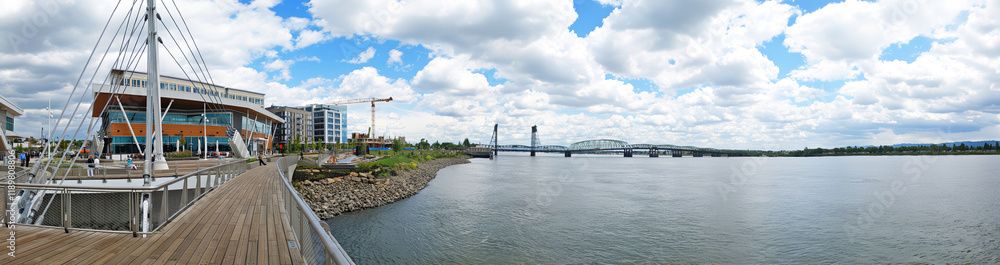 Panoramic view of Portland waterfront along the Willamette River with the Hawthorne Bridge and modern buildings under cloudy skies.