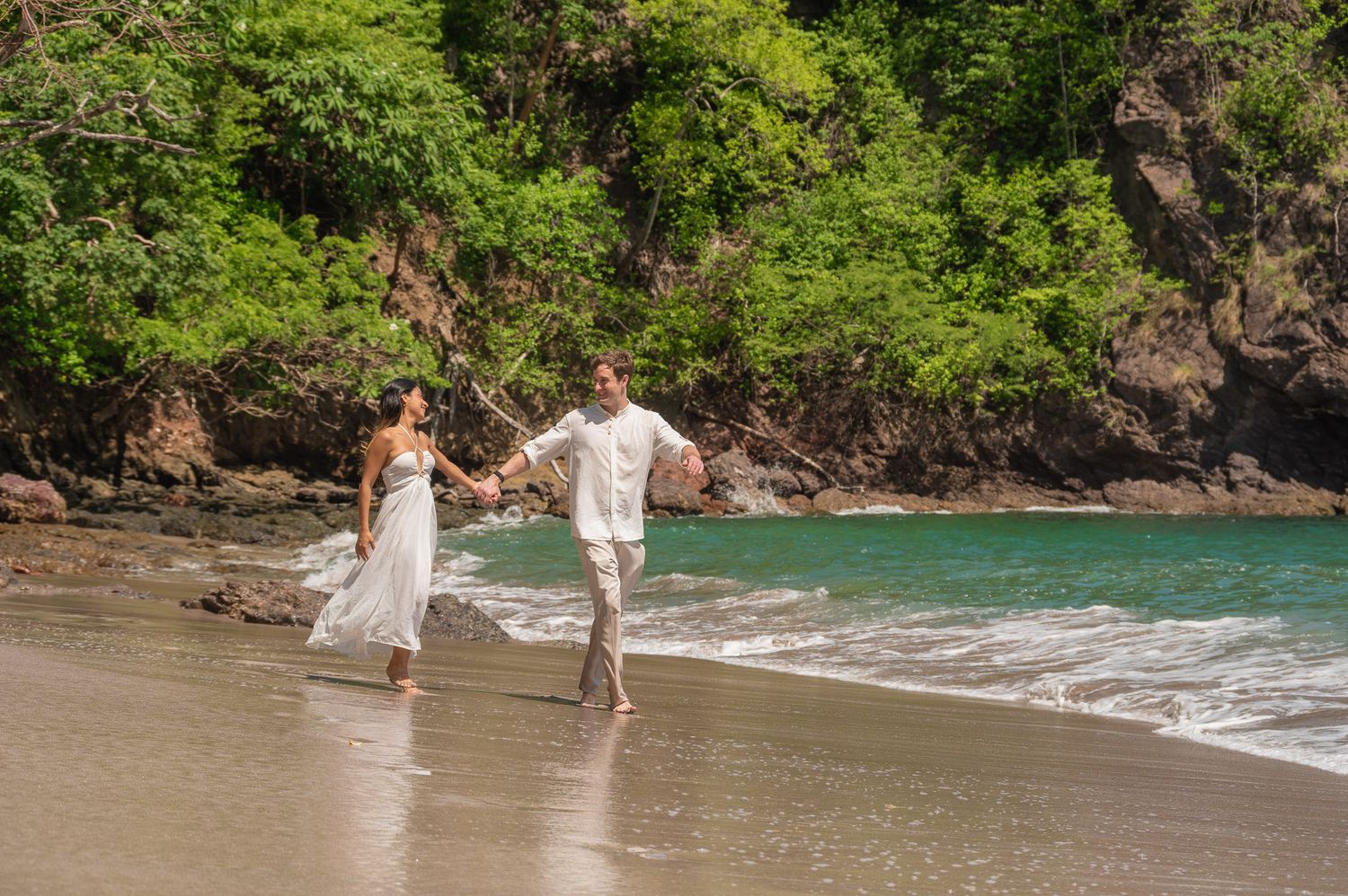 Couple in white outfits holding hands walking along a tropical beach with lush green trees and turquoise water.