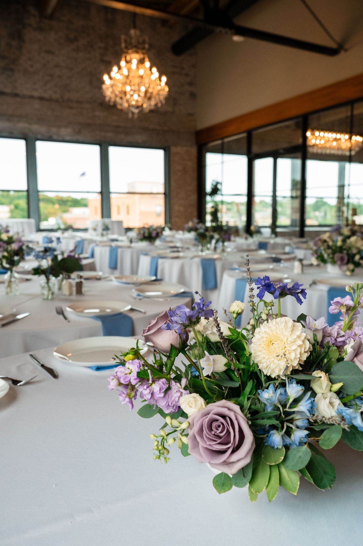 Elegant wedding reception hall with floral centerpieces featuring lavender roses, white dahlias, and blue blooms on white-draped tables.