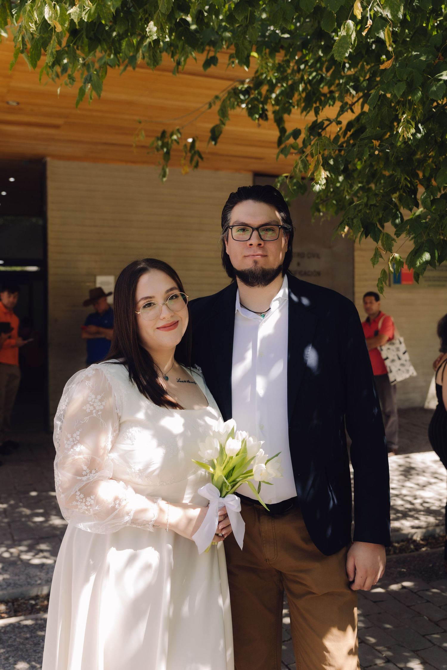 Bride holding white tulip bouquet stands with groom outside building under green tree in sunlight.