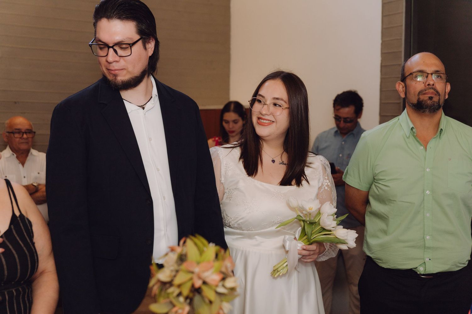 Couple stands at civil wedding ceremony indoors with guests and Chilean flag visible in background.