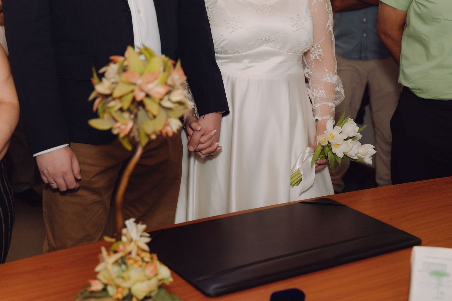 Bride and groom hold hands at civil wedding ceremony table with floral arrangement in foreground.
