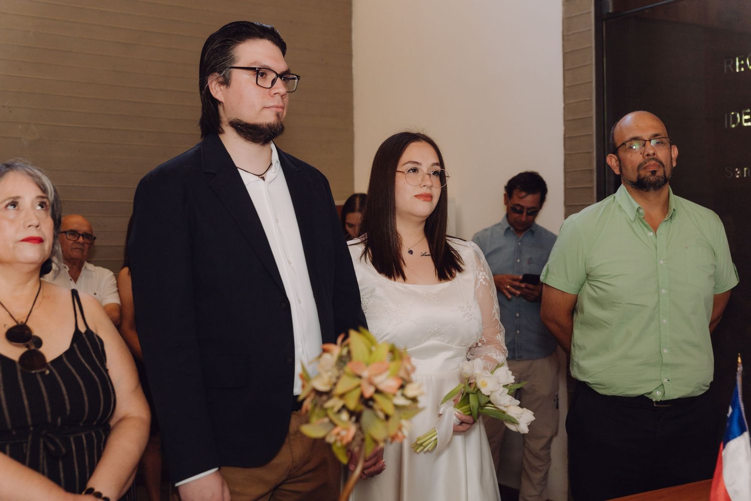 Bride holding bouquet stands beside groom and witness during indoor civil wedding ceremony.