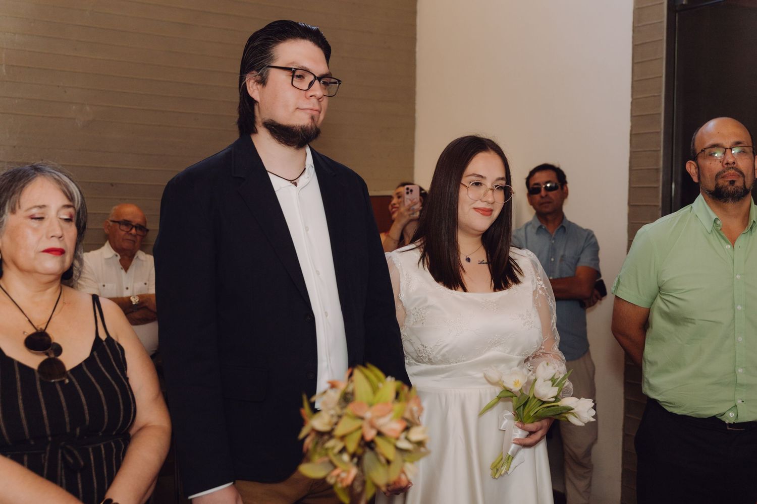 Couple stands attentively during indoor civil wedding ceremony surrounded by family and witnesses.