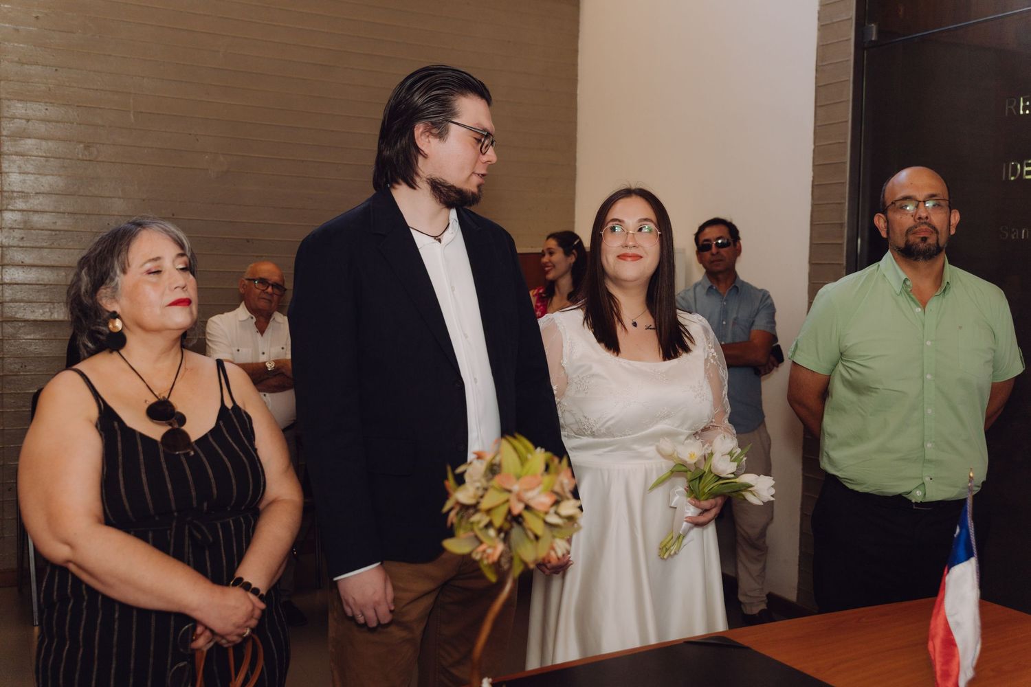 Couple stands at civil wedding ceremony with guests and Chilean flag in background.