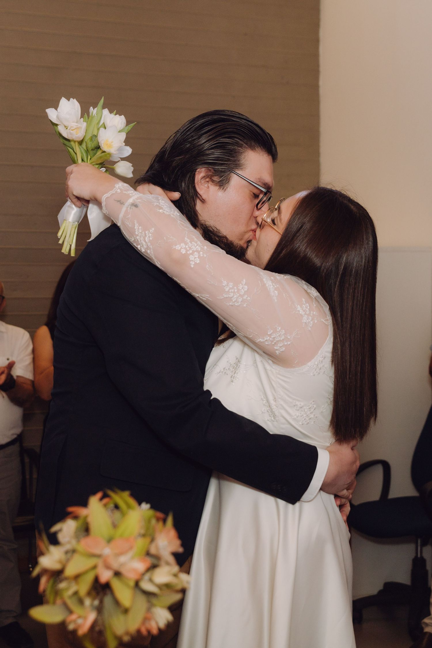 Newlyweds share first kiss as bride holds white flower bouquet at wedding ceremony.