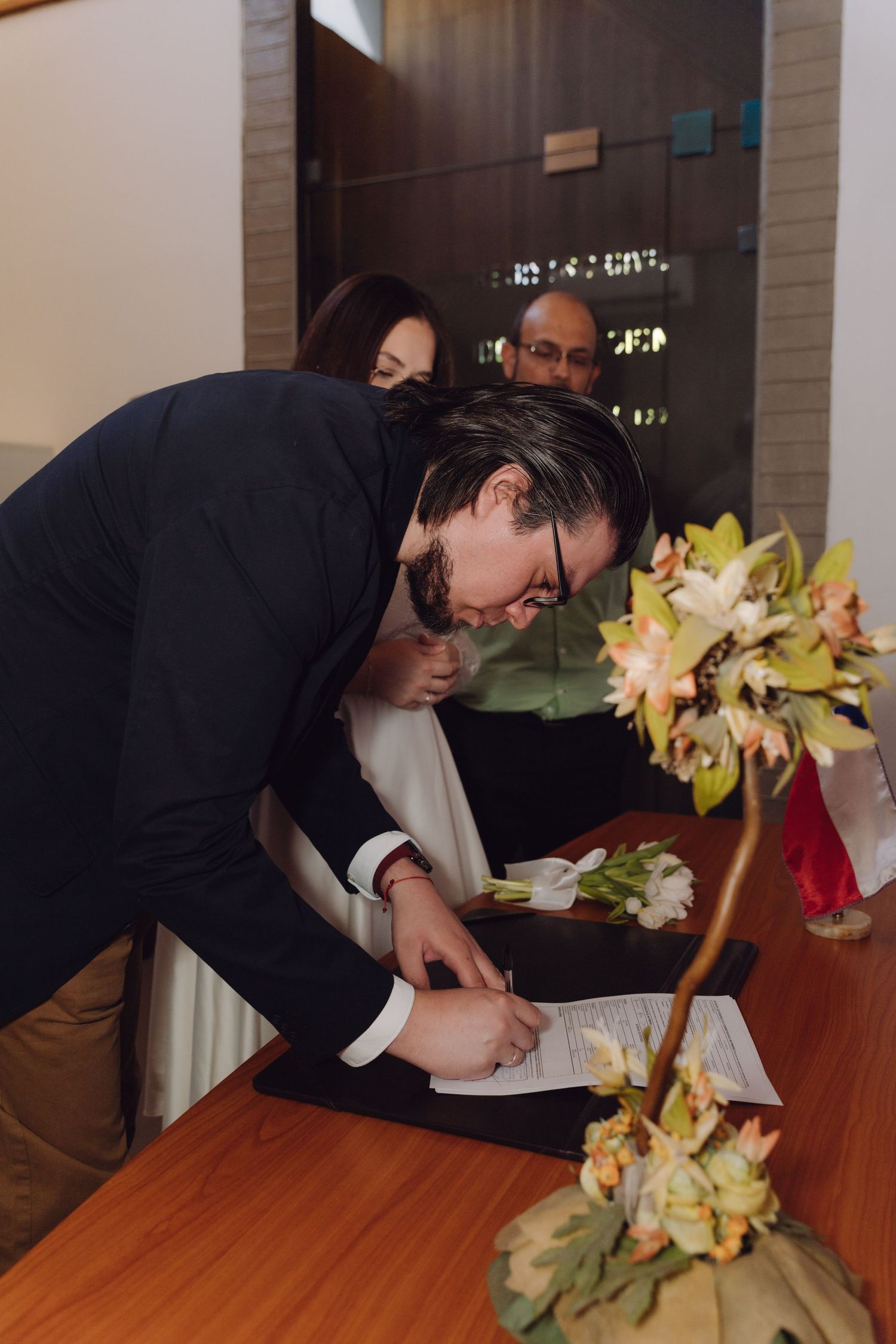 Groom signs wedding certificate at wooden table with floral arrangement nearby.
