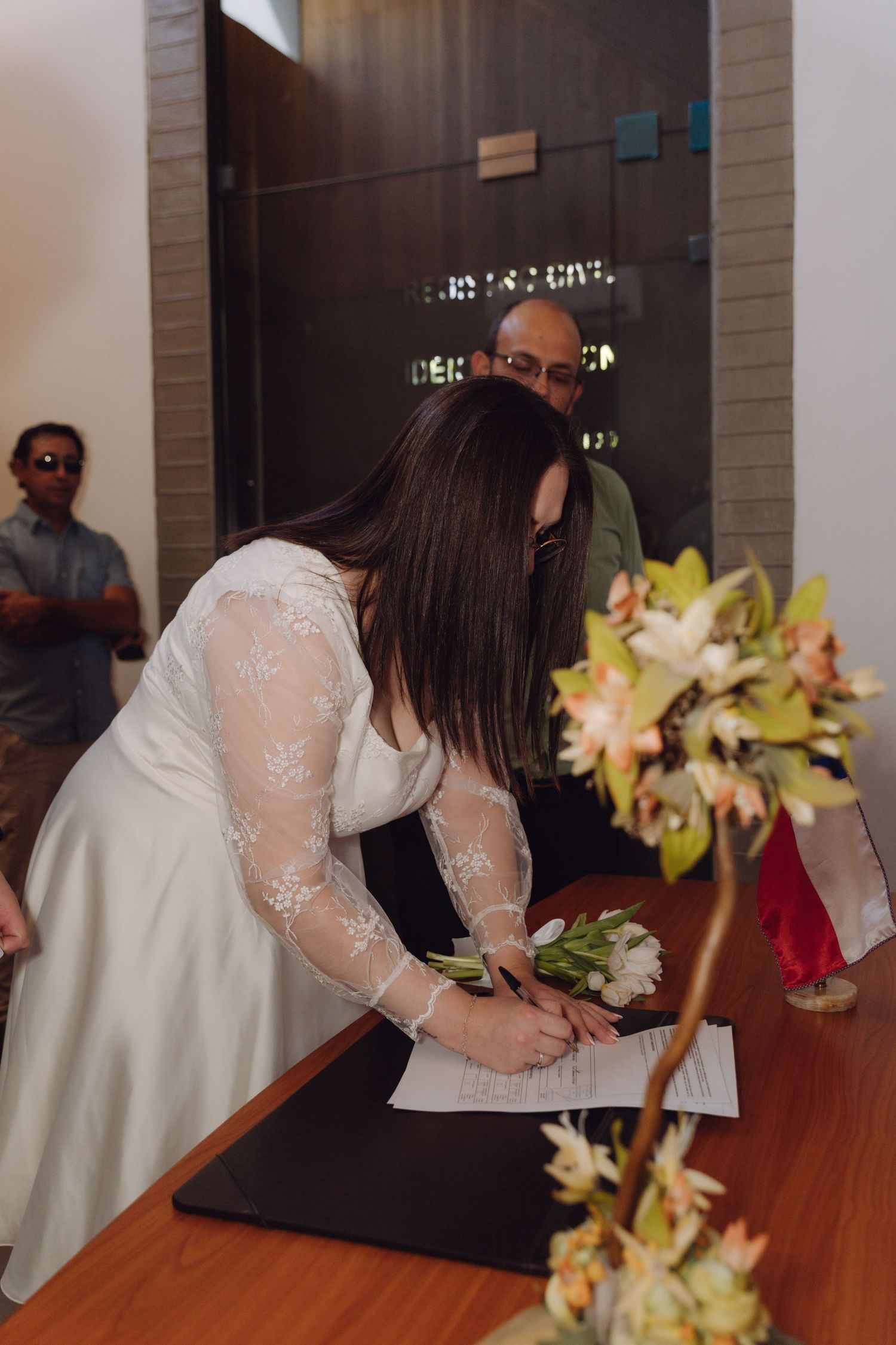 Bride signs official wedding documents at civil ceremony with guests watching.