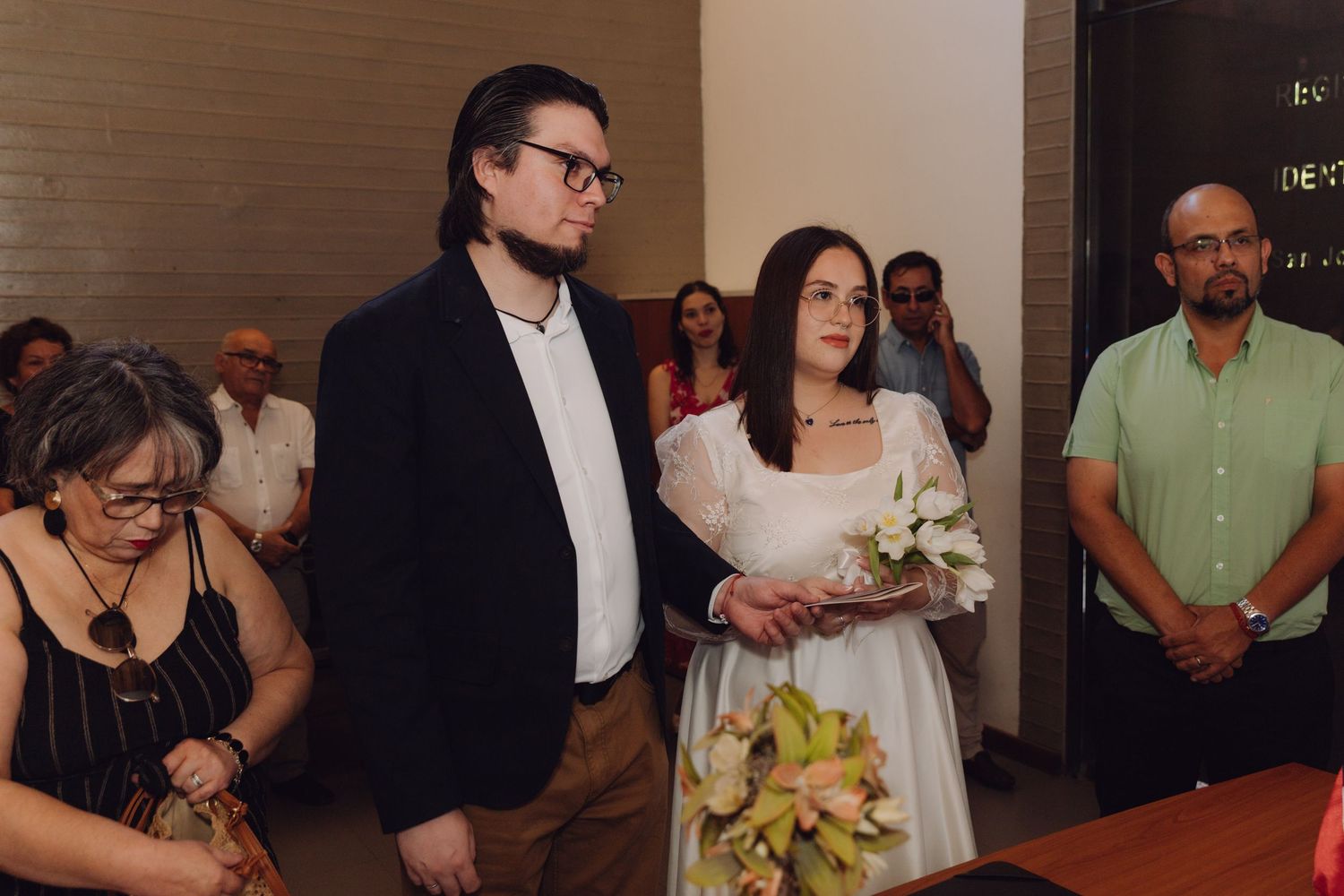 Couple stands together holding bouquet during civil wedding ceremony with guests.