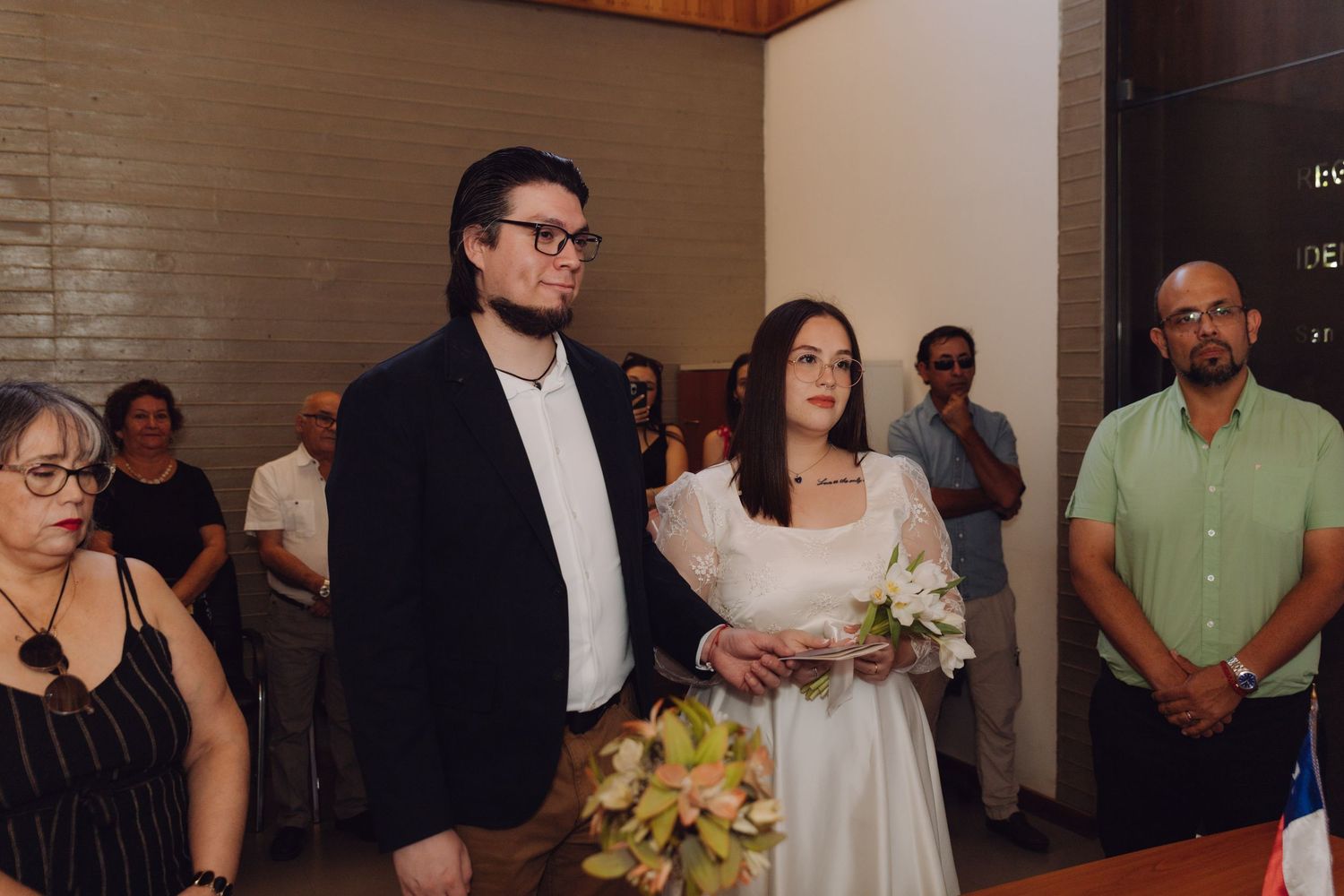Bride in white dress holds bouquet beside groom in black blazer during civil wedding ceremony with guests.