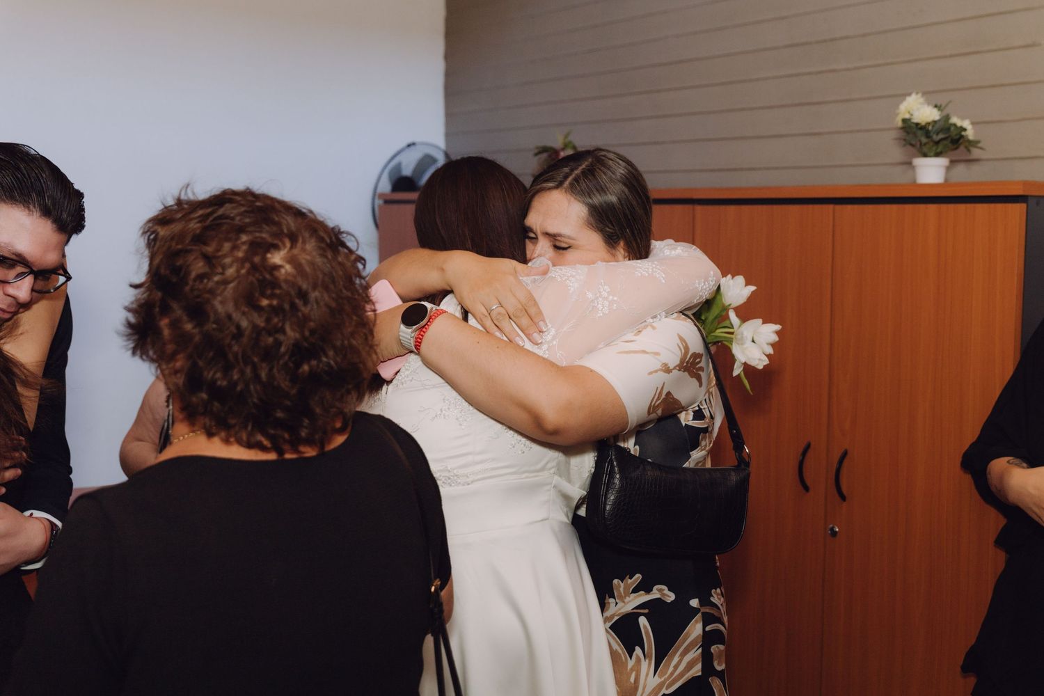 Bride in white lace dress receives warm hug from guest while holding white flowers at wedding reception.