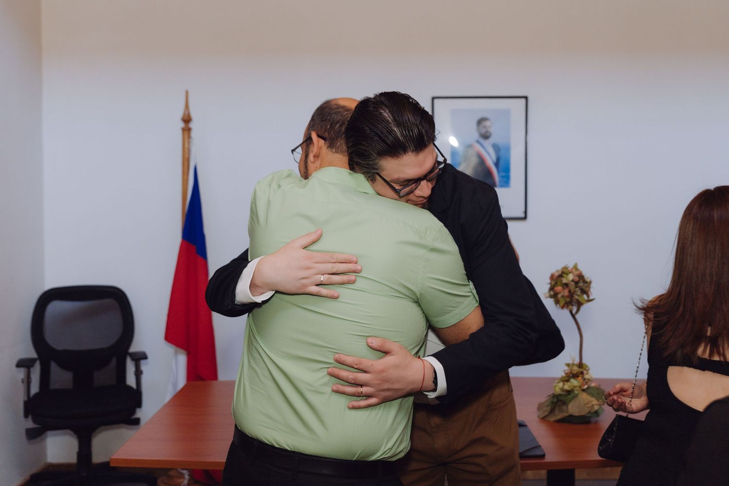 Two men share an emotional hug near a Chilean flag during a wedding celebration indoors.