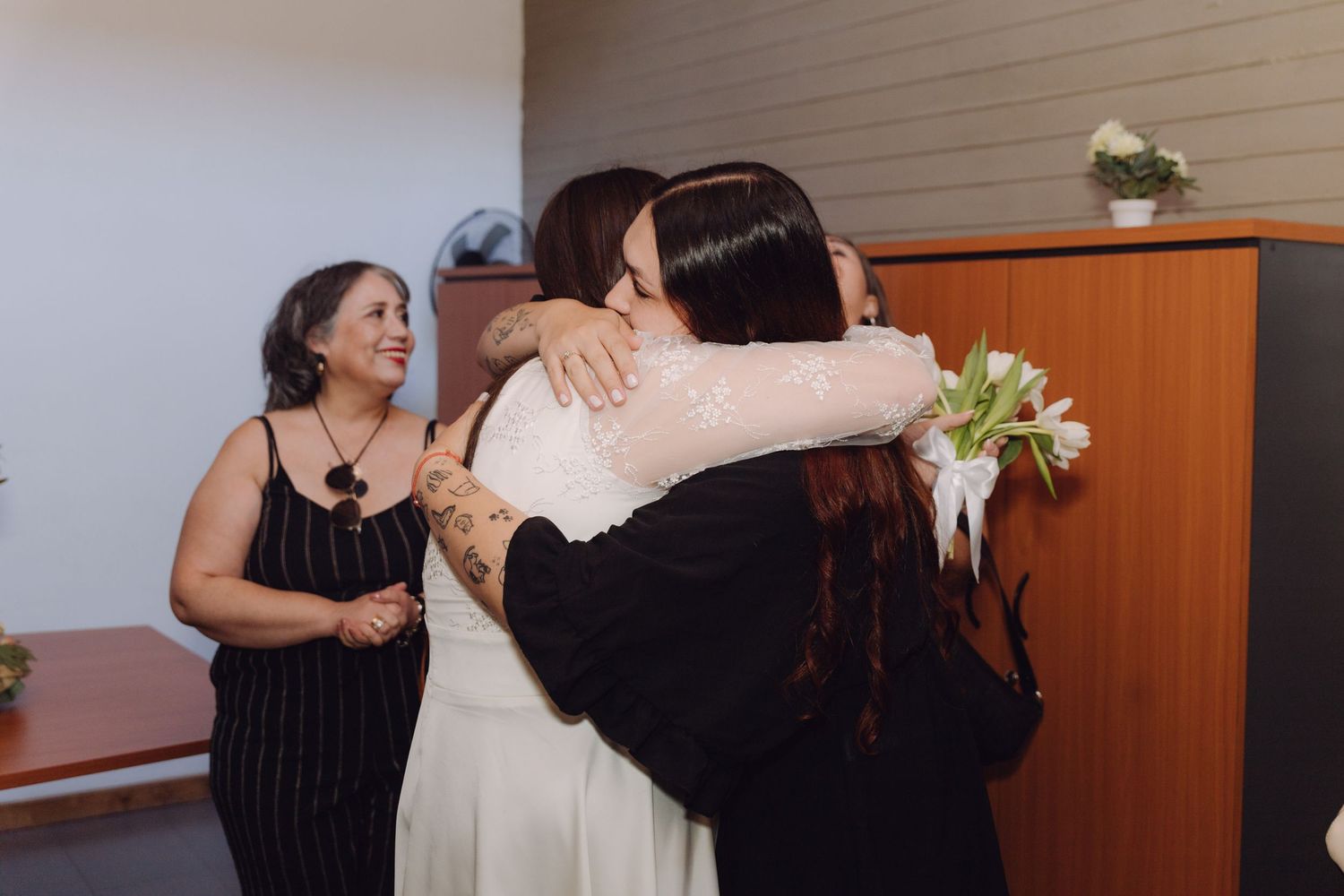 Bride in white lace dress hugs a guest warmly while holding white tulips at wedding reception.