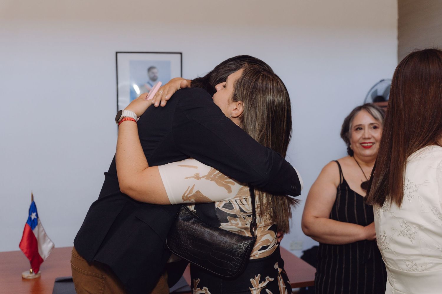 Two women embrace warmly at a wedding celebration with Chilean flag and guests visible in background.