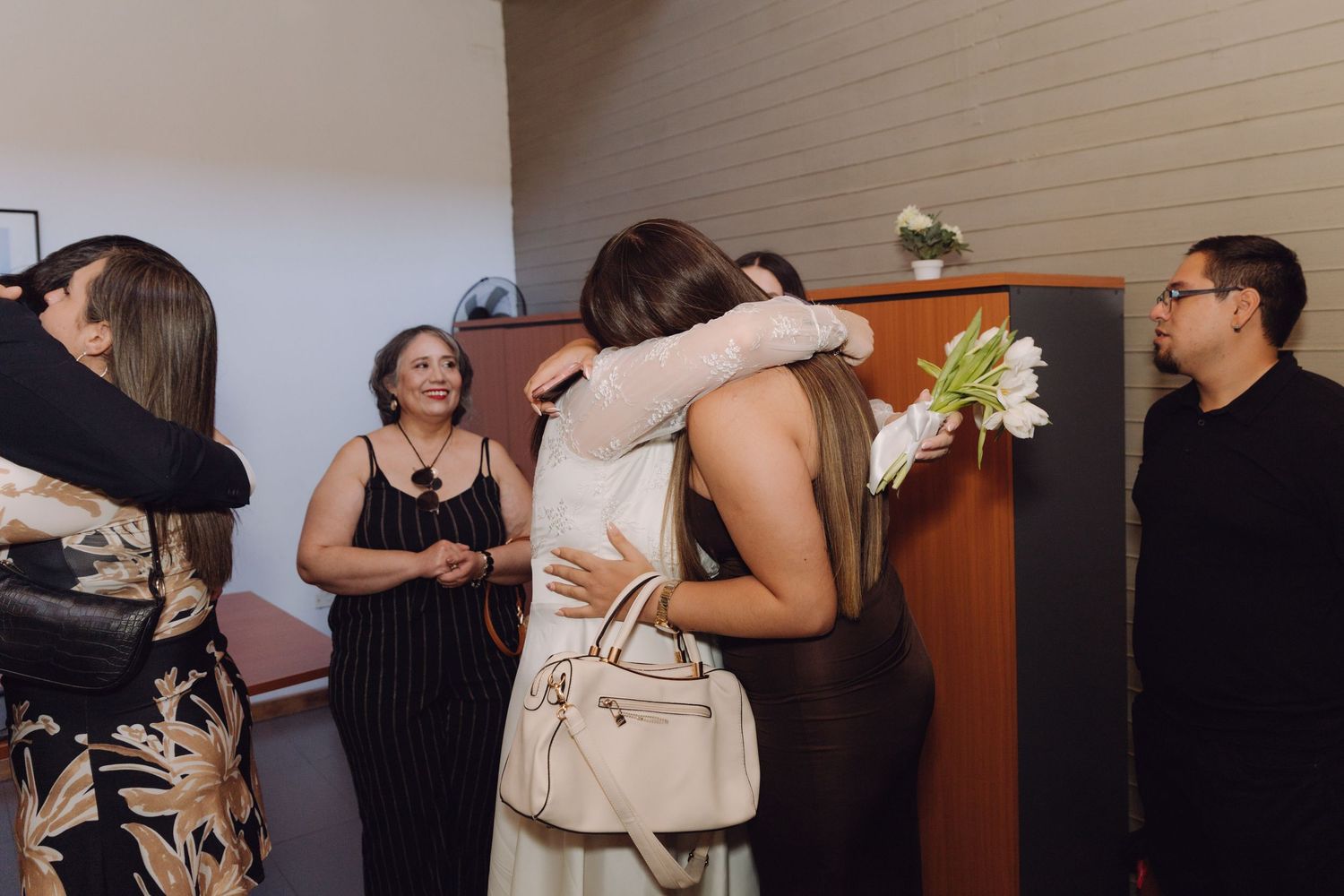 Bride holding white tulips receives congratulatory hugs from guests at intimate wedding celebration indoors.