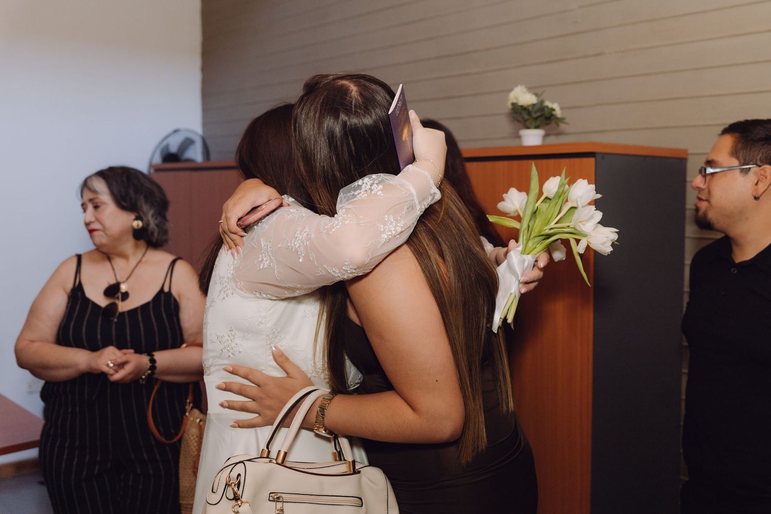 Bride in white lace dress embraces guest while holding white tulips at joyful wedding reception.