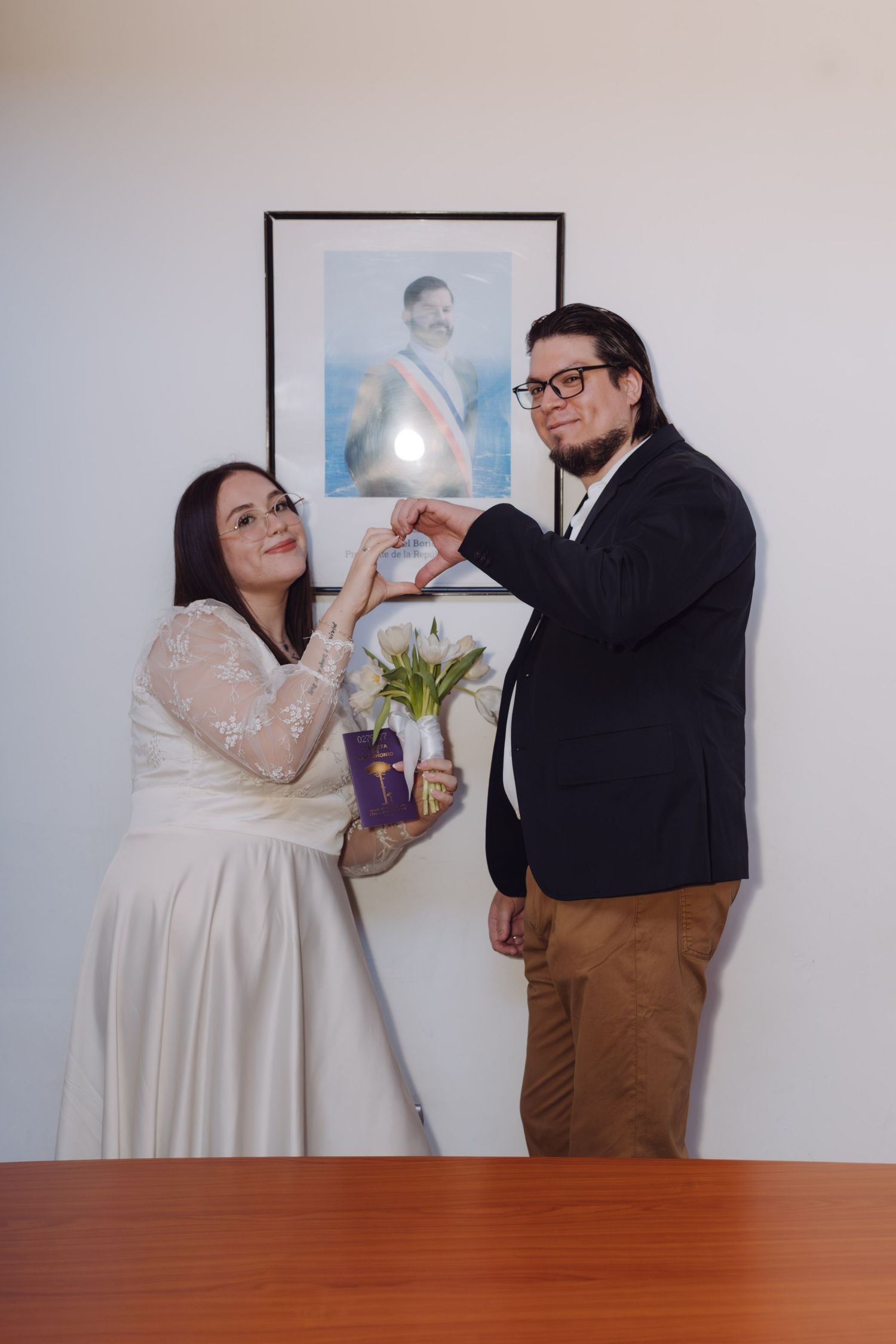Newlyweds make heart shape with hands holding white tulips and gift in front of portrait.