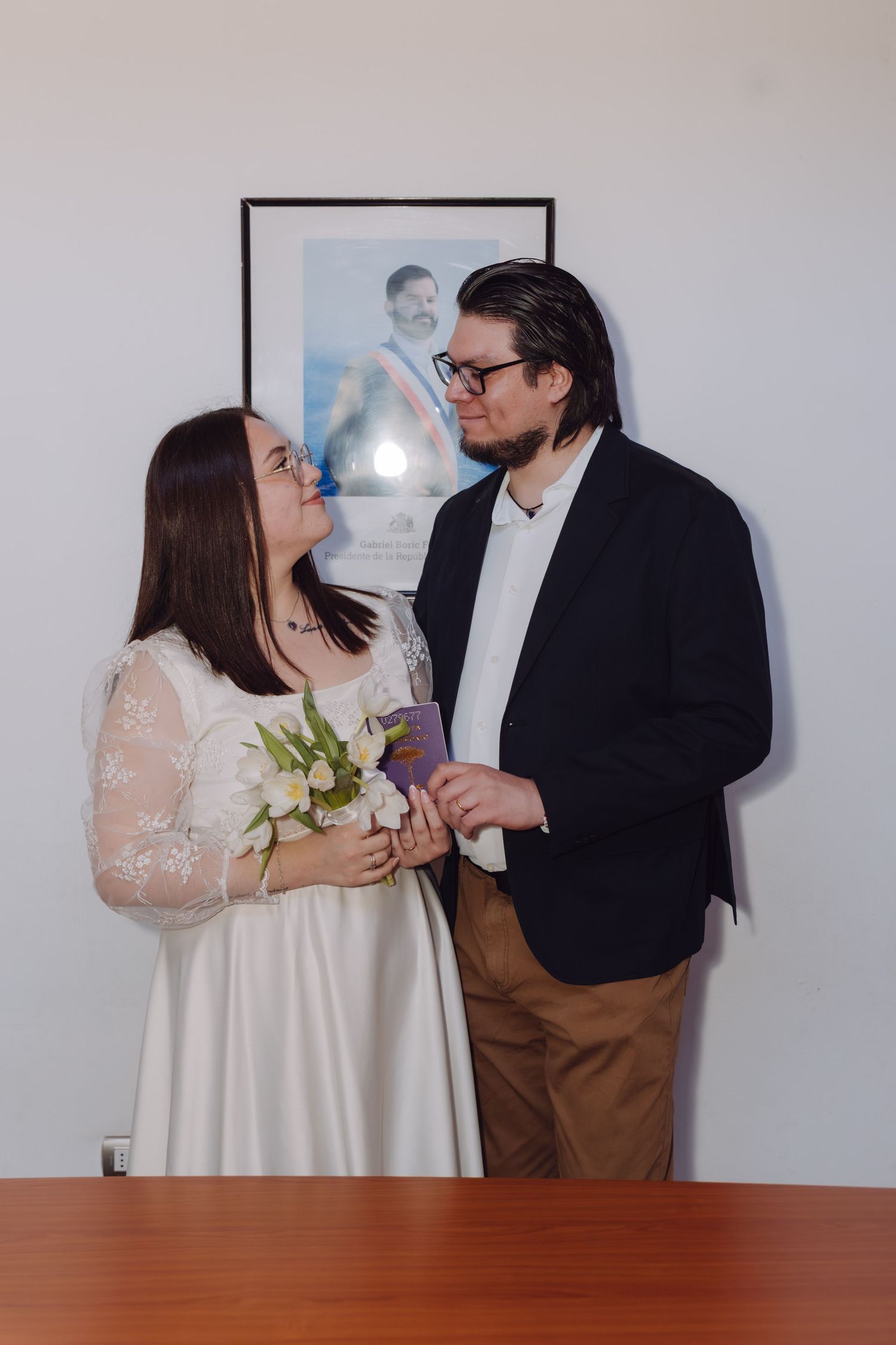 Newlywed couple gazes lovingly at each other holding flowers and documents at civil wedding ceremony.