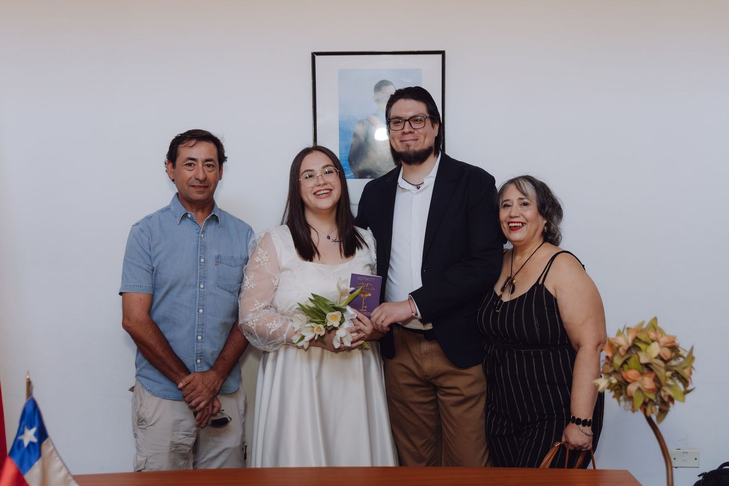 Wedding couple stands with two family members smiling at a civil ceremony with Chilean flag visible.