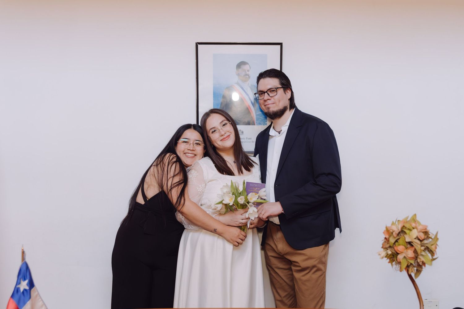 Bride embraces a female guest while groom stands beside them holding a white flower bouquet indoors.
