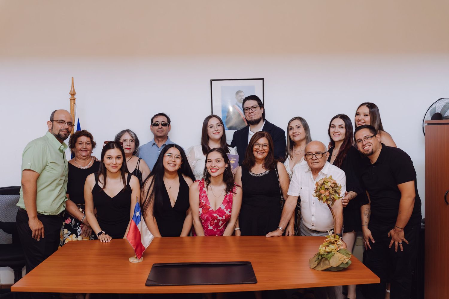 Large wedding group photo with bride and groom surrounded by family and friends at a wooden table.