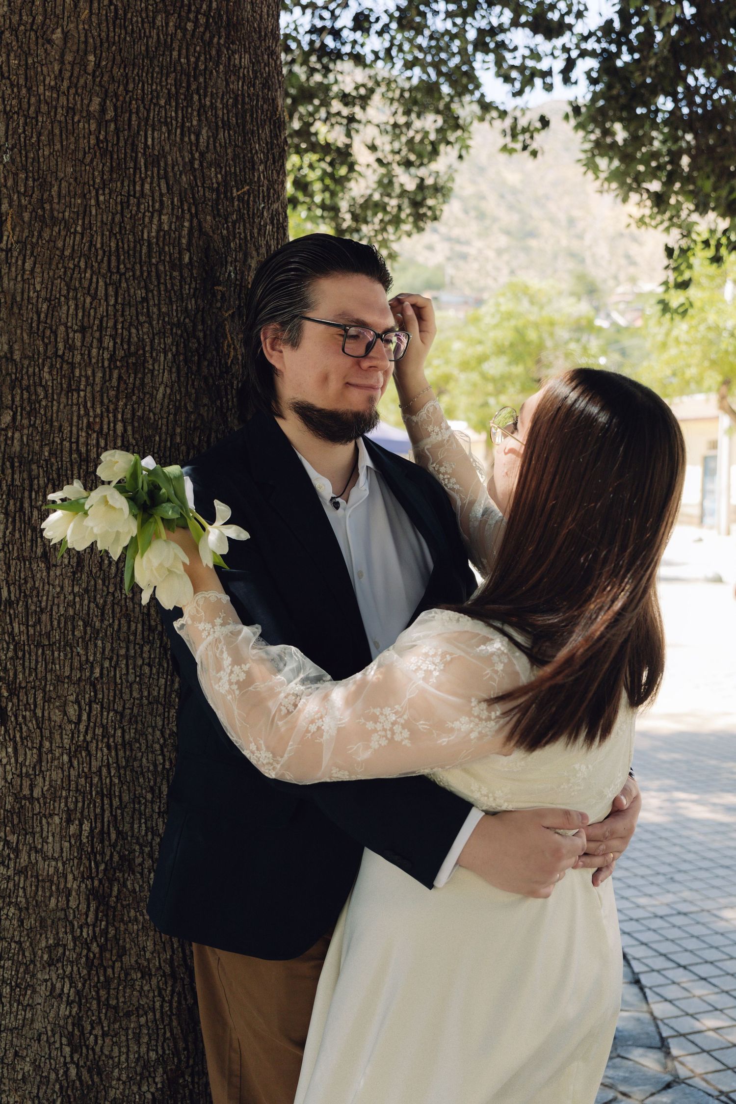 Groom in black blazer leans against tree as bride in white lace dress adjusts his hair outdoors.