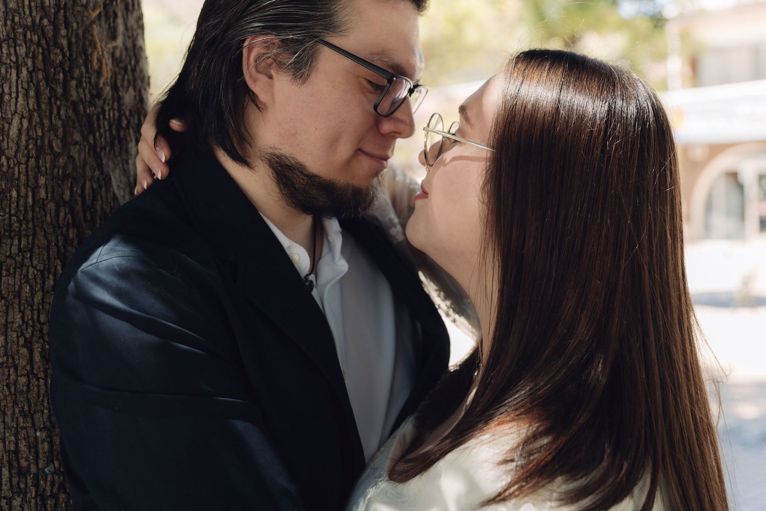 Bride and groom share an intimate almost-kiss moment leaning against a tree in sunny outdoor setting.