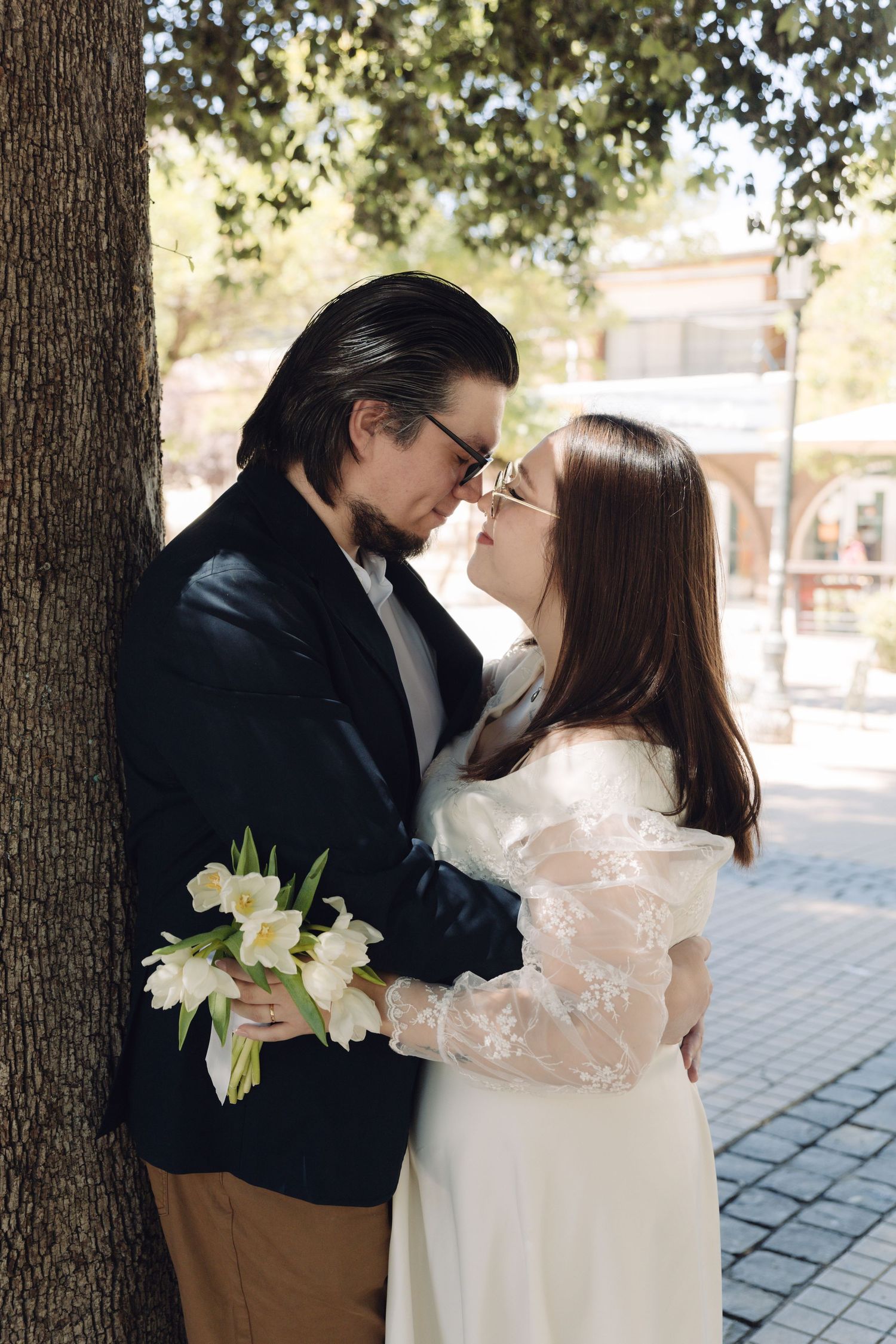 Wedding couple nearly kissing beside a tree, bride holding white tulip bouquet in sunny park setting.