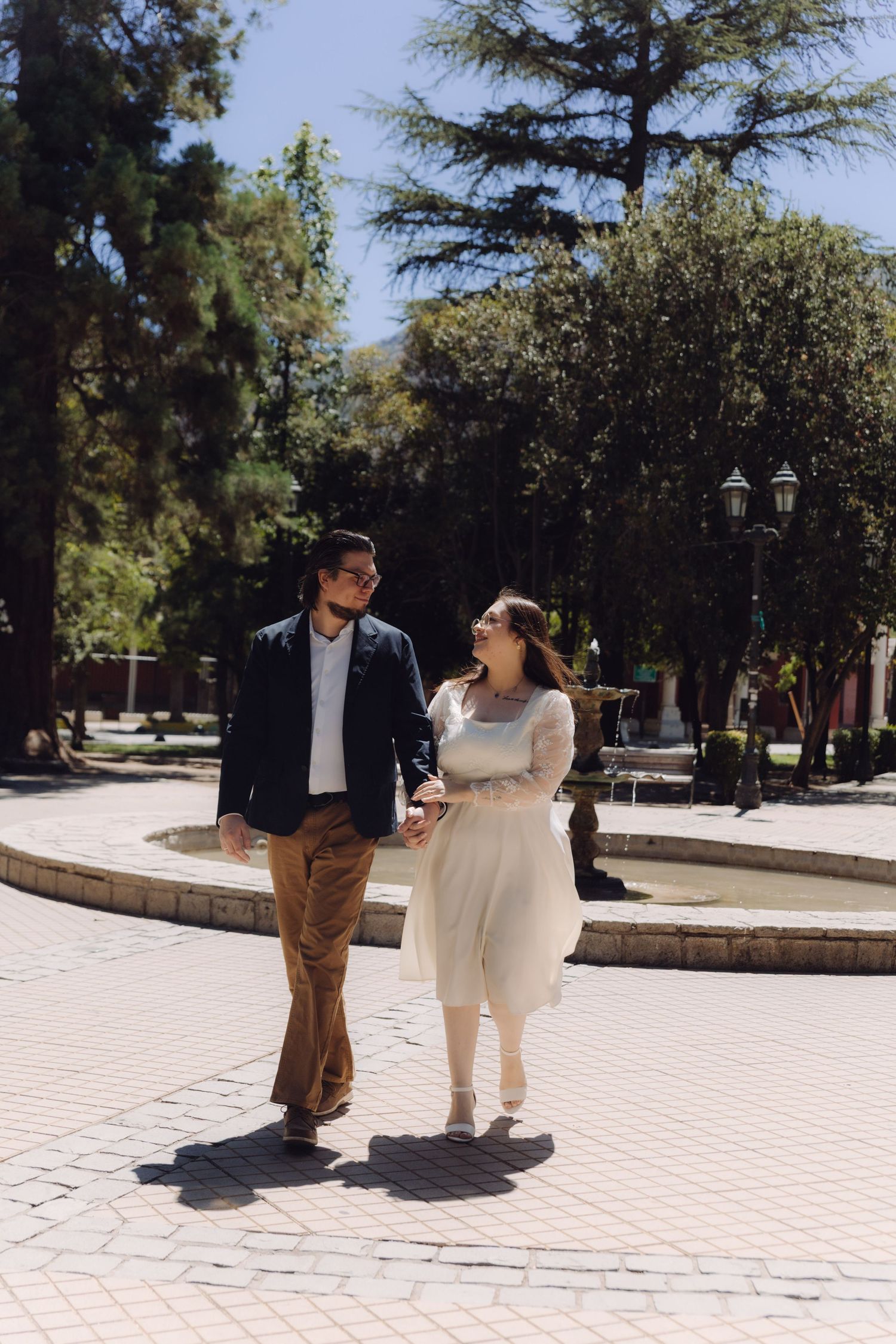 Newlywed couple walks hand in hand through a sunny plaza with fountain and trees in background.