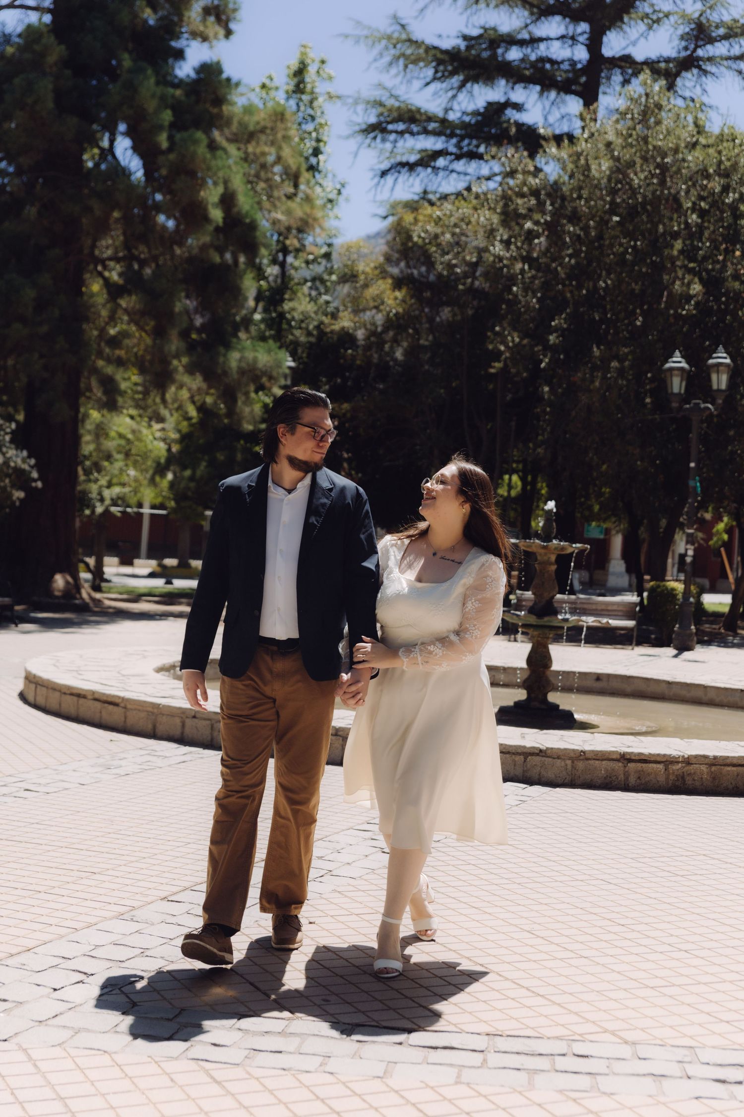 Smiling bride and groom walk hand in hand past a fountain in a sunlit outdoor plaza setting.