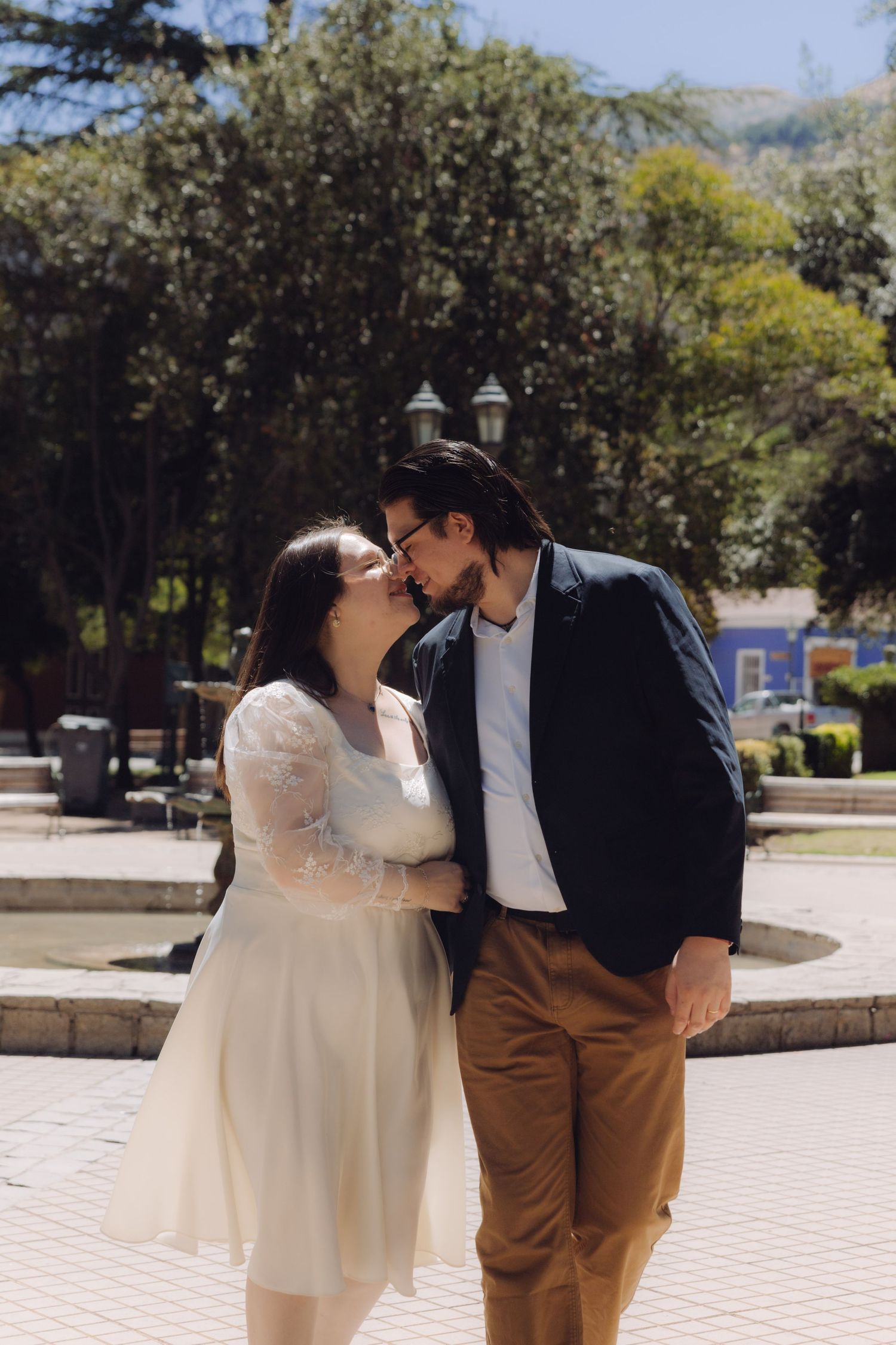 Couple in wedding attire sharing a romantic moment near a fountain in a sunny park.