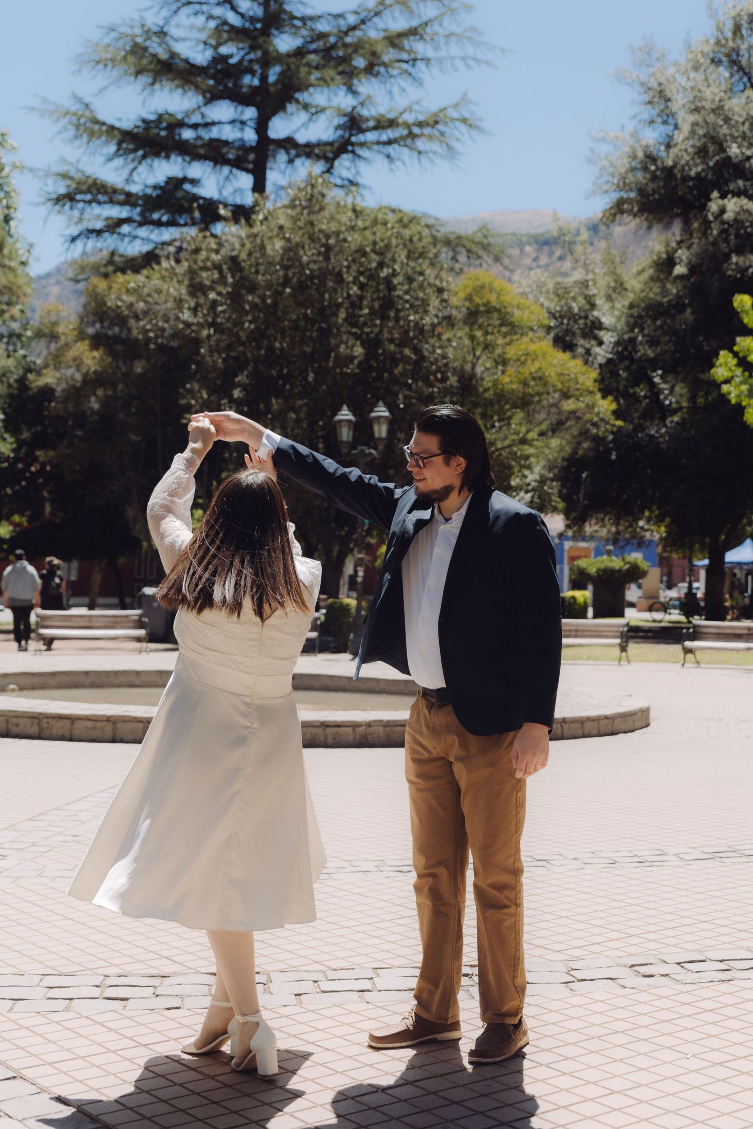 Groom spins bride in white dress in a sunny plaza with trees and fountain in the background.