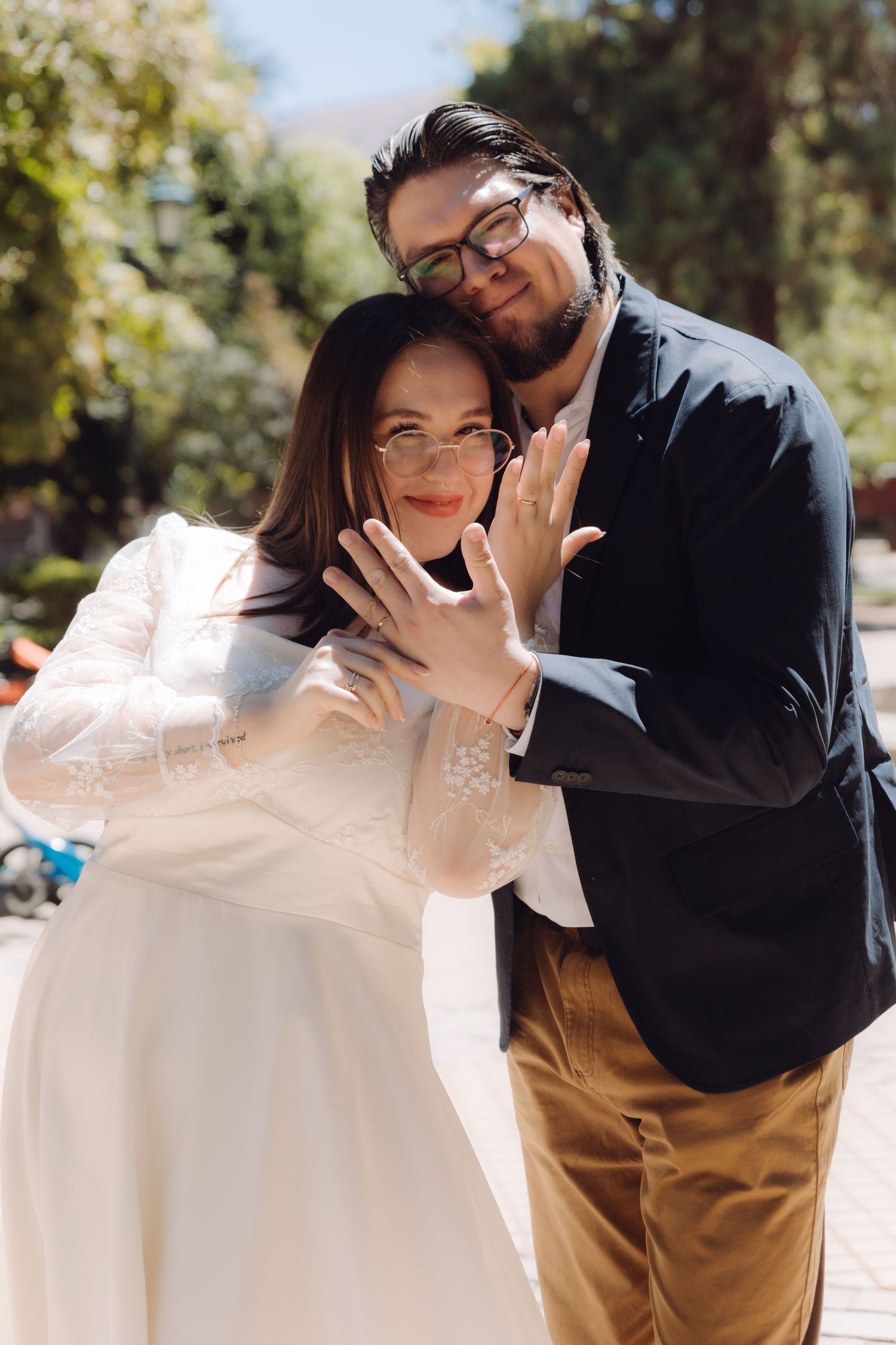 Newlyweds showing off wedding rings while embracing outdoors on a sunny day.