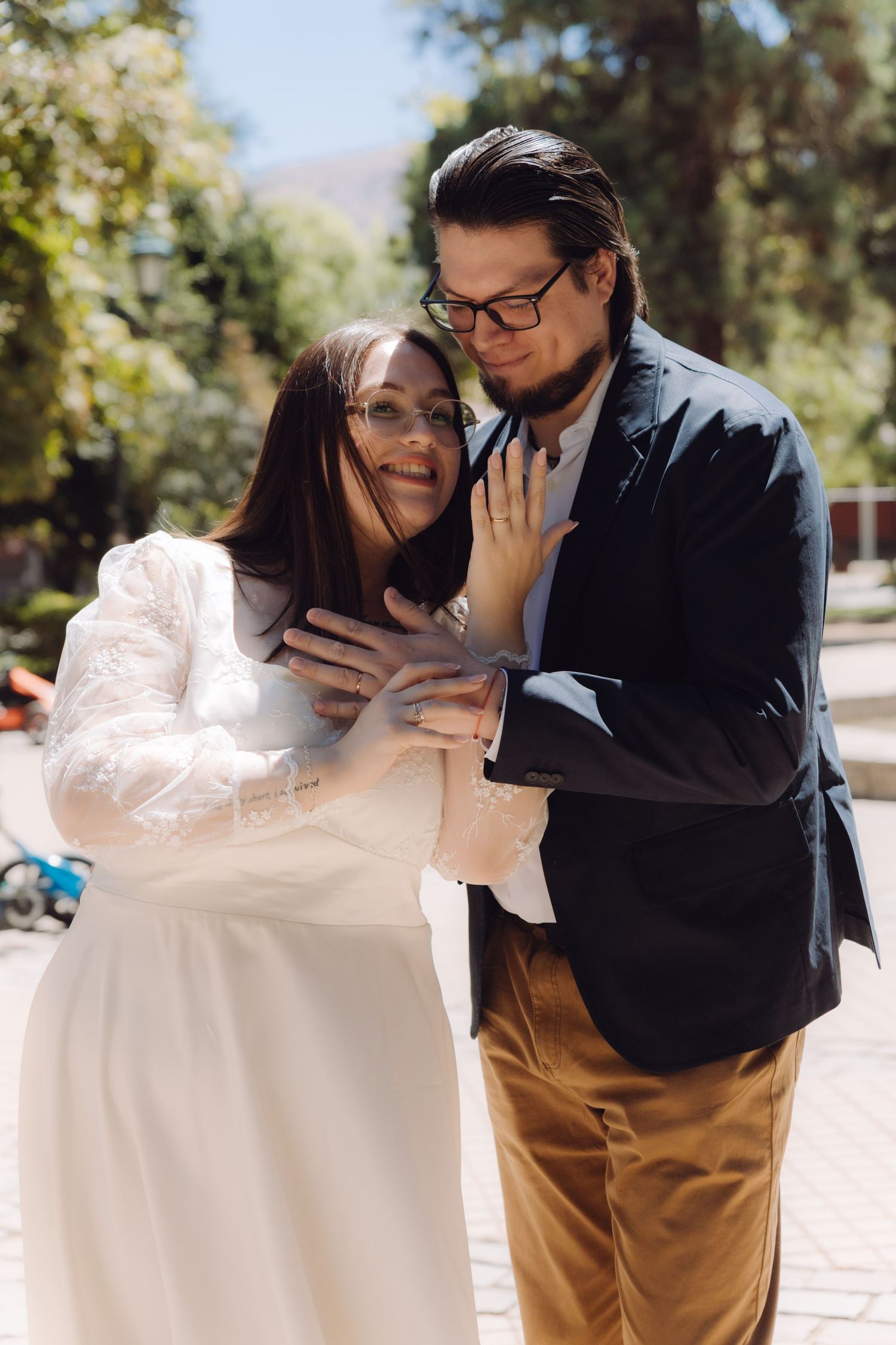 Groom in navy blazer tenderly embracing smiling bride in white lace dress outdoors.