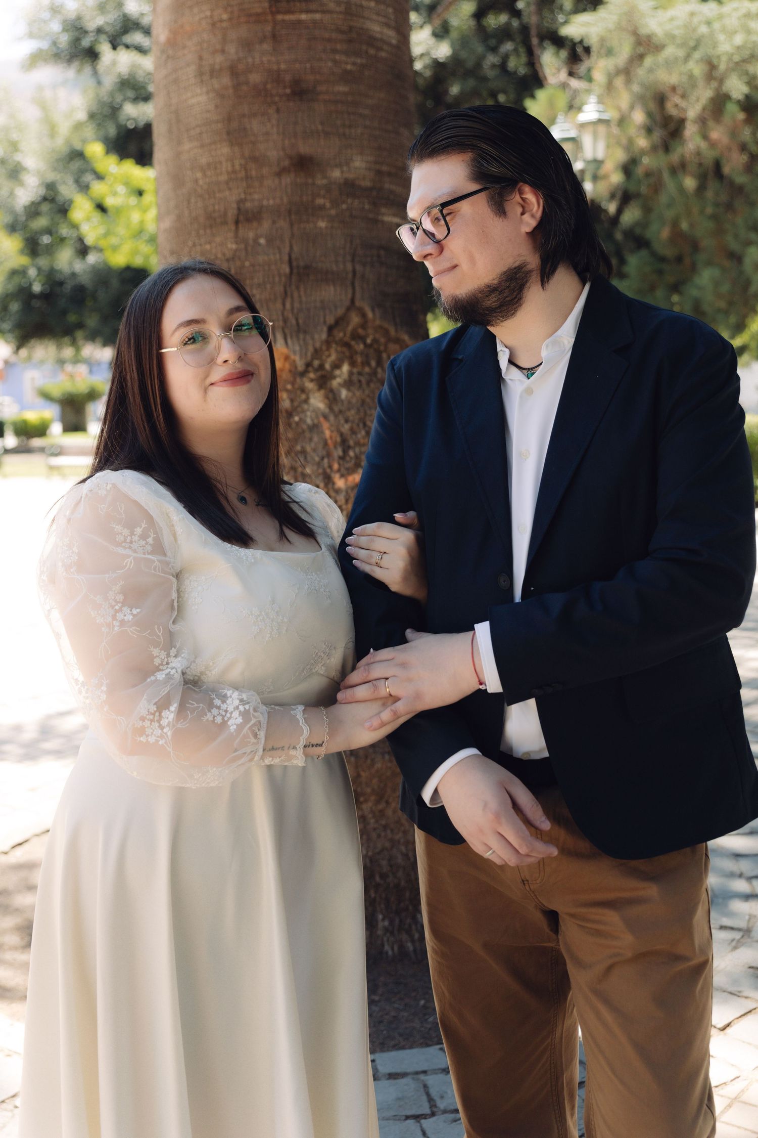 Bride and groom in elegant attire posing together beside a large tree in a park.
