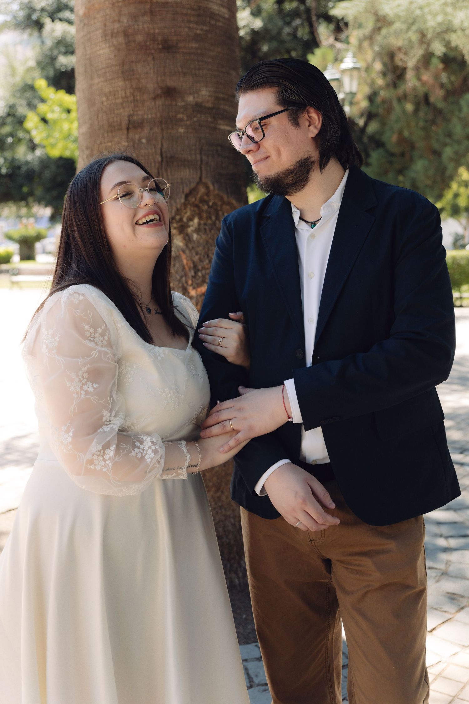 Happy couple in wedding attire laughing together beside a palm tree in sunny park.