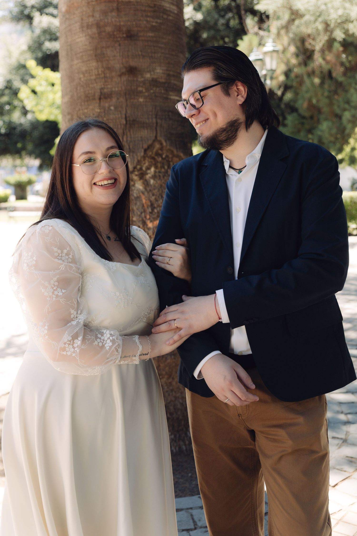 Smiling bride in white lace dress and groom in navy blazer posing by a palm tree.