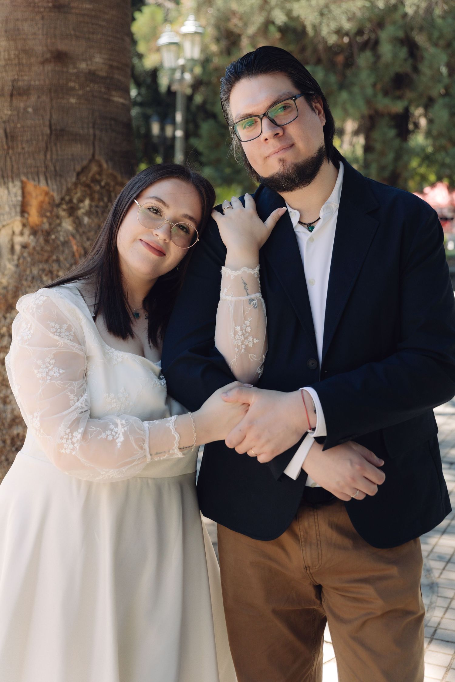 Couple in wedding attire posing together beside a palm tree in a sunlit park.