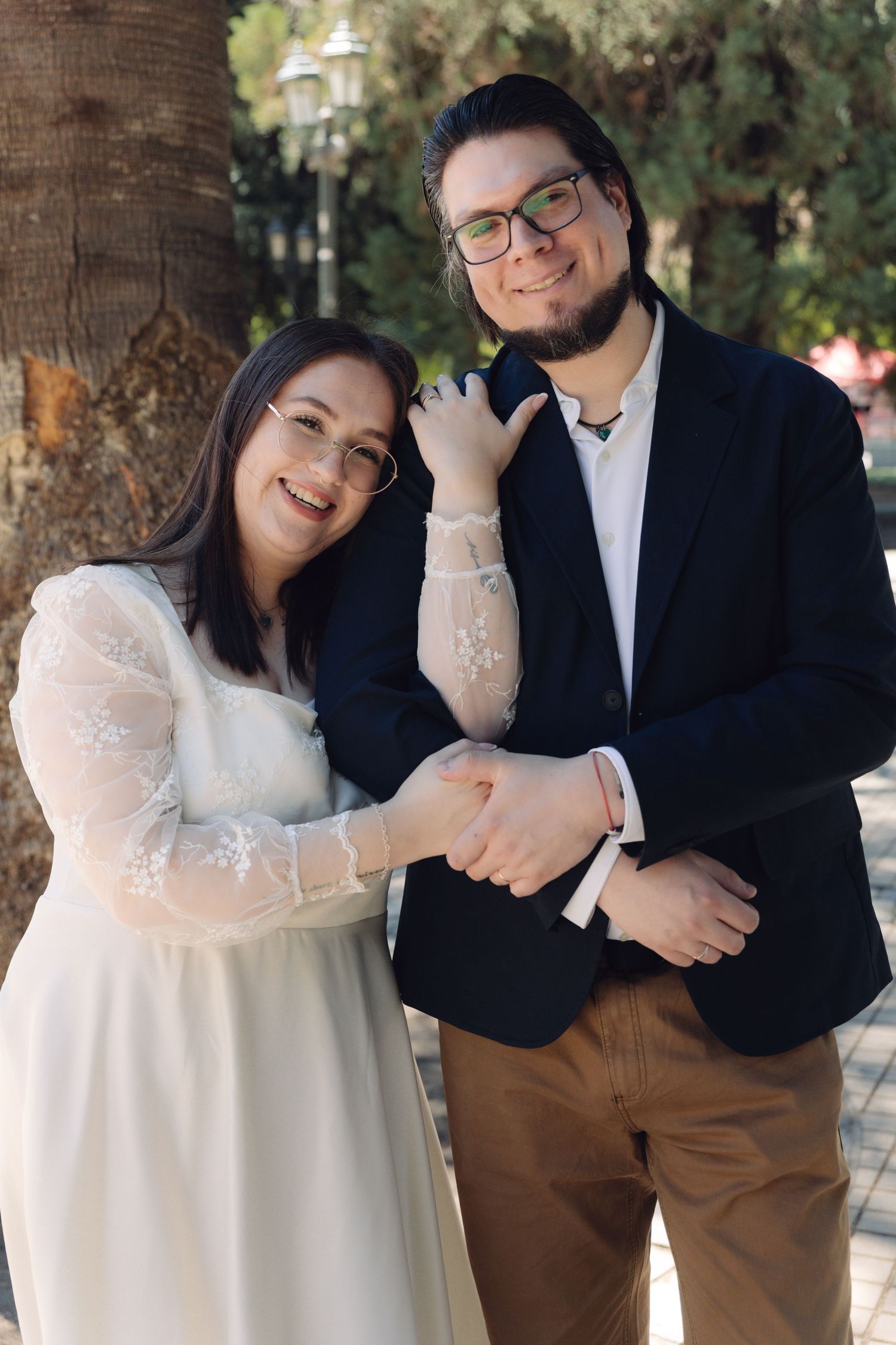 Smiling newlyweds in elegant wedding attire posing happily beside a park palm tree.