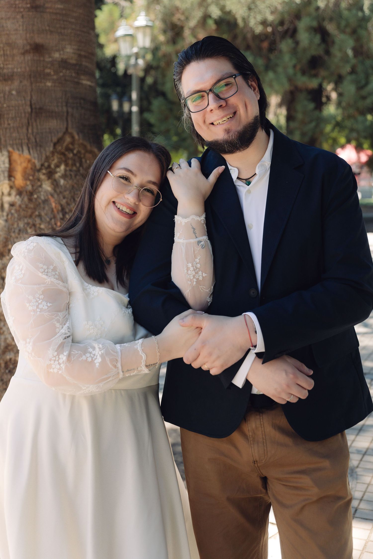 Smiling couple in wedding attire poses outdoors, bride in white lace dress and groom in dark blazer with glasses.