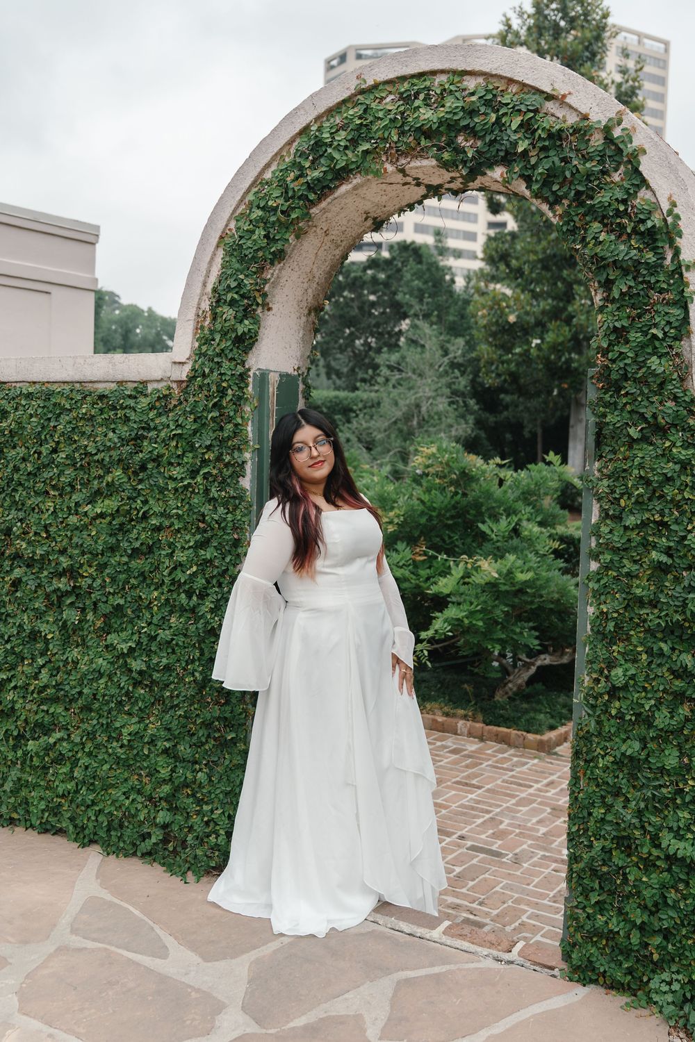 Bride in white bell-sleeve gown standing under ivy-covered stone arch in a lush garden setting.