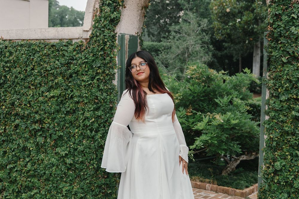 Woman in white bell-sleeve gown poses outdoors among lush green ivy-covered walls, wearing glasses.