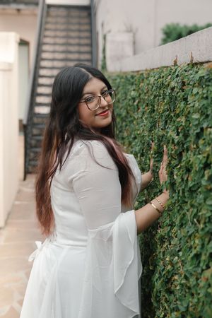 Young woman in white dress and glasses smiling beside a lush green ivy wall with stairs in background.